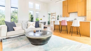 A spacious living area with a large round coffee table, a sofa with cushions, and a kitchen area with wooden cabinets and bar stools. - Glamorous Perfectly Located Deluxe 2 Bed Townhome (Dallas)