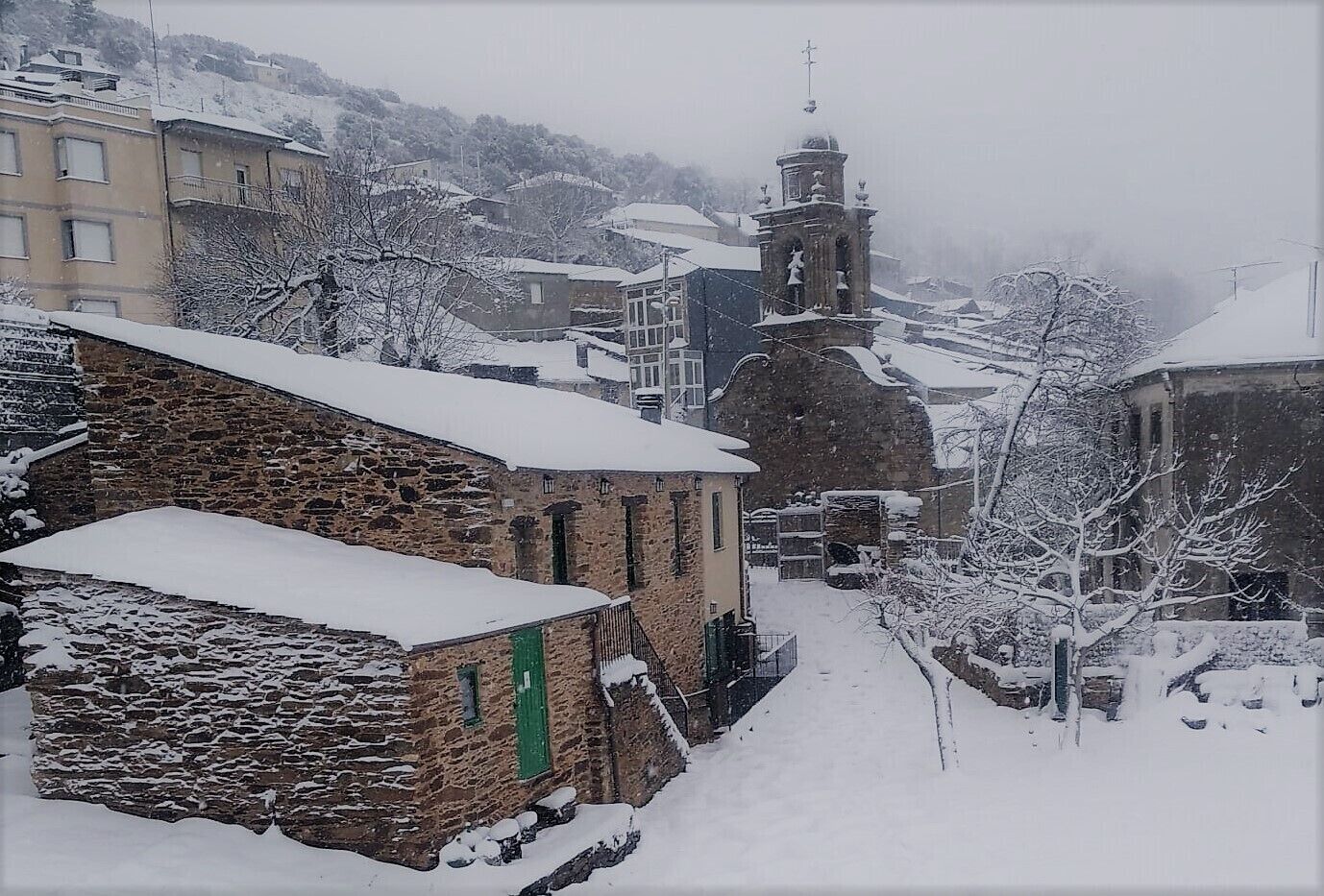 Un poble cobert de neu amb edificis de pedra, una església amb un campanar i un carrer estret.