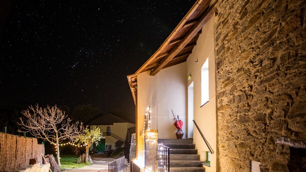 A stone building with a wooden staircase leading to a door, a tree with lights, and a starry night sky. - Eco dos Teixos (Carballeda de Valdeorras)