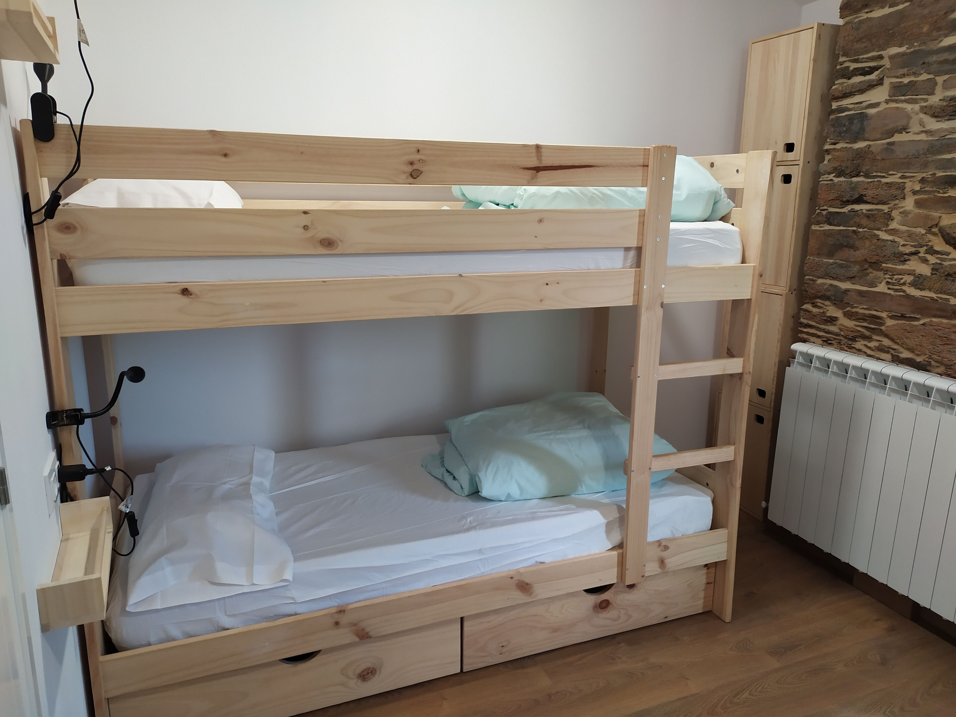 En køjeseng med træramme, en hvid madras og en lyseblå pude. Der er en hvid radiator på gulvet ved siden af sengen.