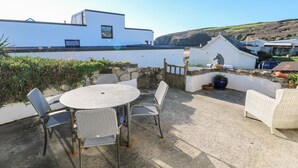 Eine Terrasse mit rundem Tisch und Stühlen, einer Steinmauer und Blick auf ein weißes Gebäude sowie Hügel im Hintergrund.