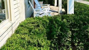 A covered porch with white chairs and a small table, surrounded by well-maintained bushes.