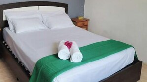 Ein ordentlich gemachtes Bett mit weißen Leinen und einer grünen Tagesdecke, ein Nachttisch aus Holz mit einer Lampe und eine graue Wand.