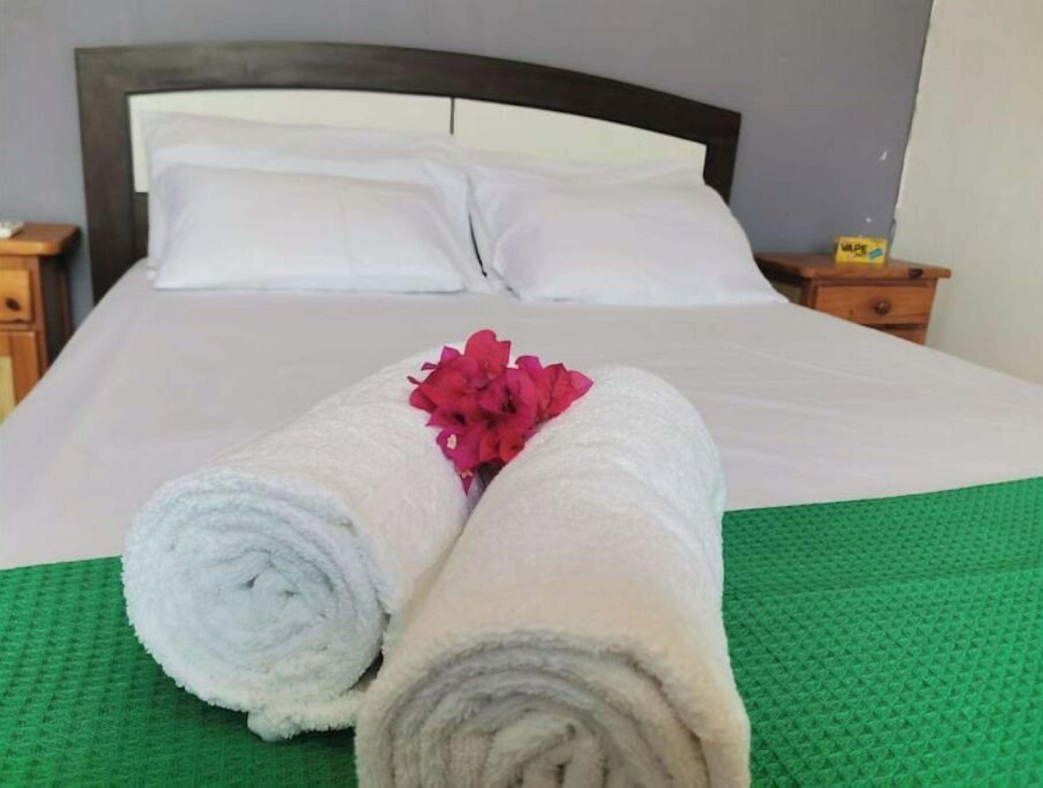 Ein ordentlich gemachteres Bett mit weißen Leinen und einer grünen Tagesdecke, ergänzt durch zwei zusammengerollte Handtücher und eine rosa Blume.