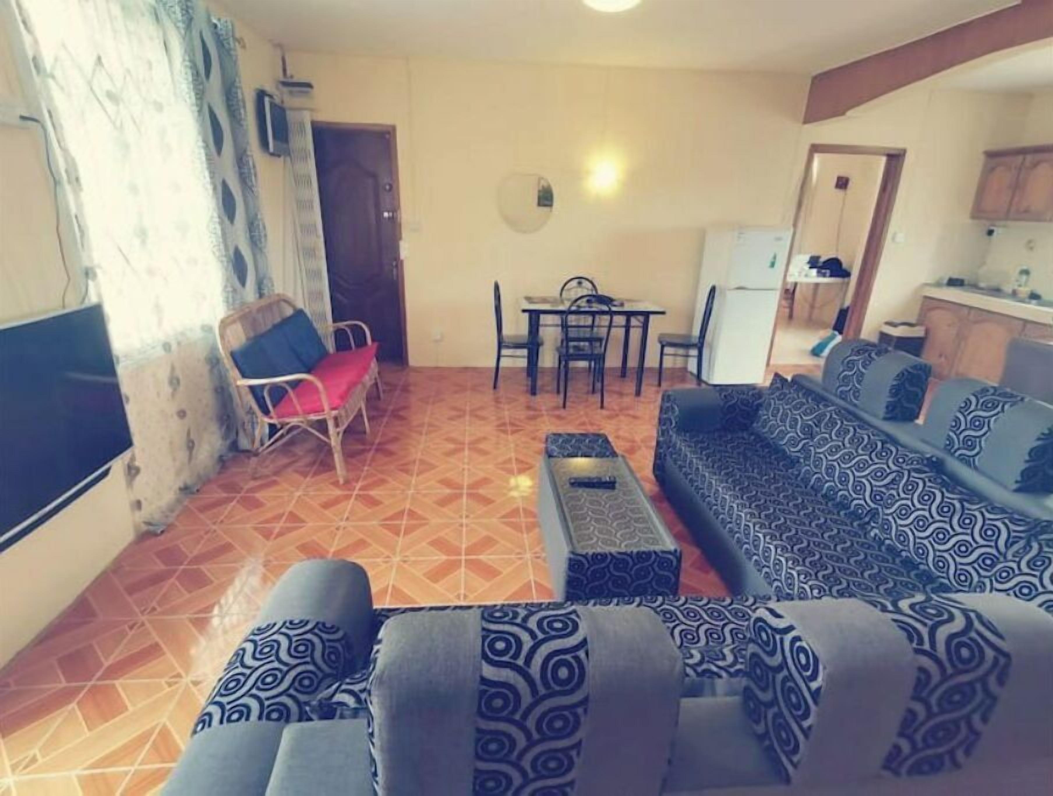 A living room with a sofa, a TV, a dining table with chairs, and a kitchen area in the background.