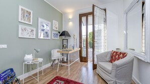 Una habitación con escritorio, silla y sofá. Hay cuadros en la pared y una ventana con cortinas.