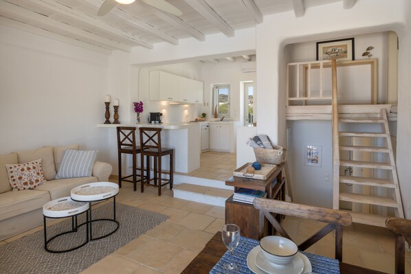 A cozy living room with a sofa, two side tables, and a coffee table. A kitchen is visible in the background with white cabinets and a wooden ladder. - Naousa Bay Views with pool - Villa Caroline (Naousa)