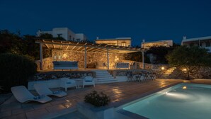 Un espace aménagé au bord de la piscine, avec des chaises longues, un mur en pierre et une pergola dotée d’un éclairage.