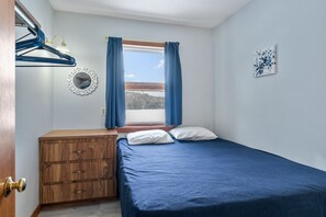 A bedroom with a wooden dresser, a bed with blue bedding, a window with curtains, a wall-mounted mirror, and a small artwork.