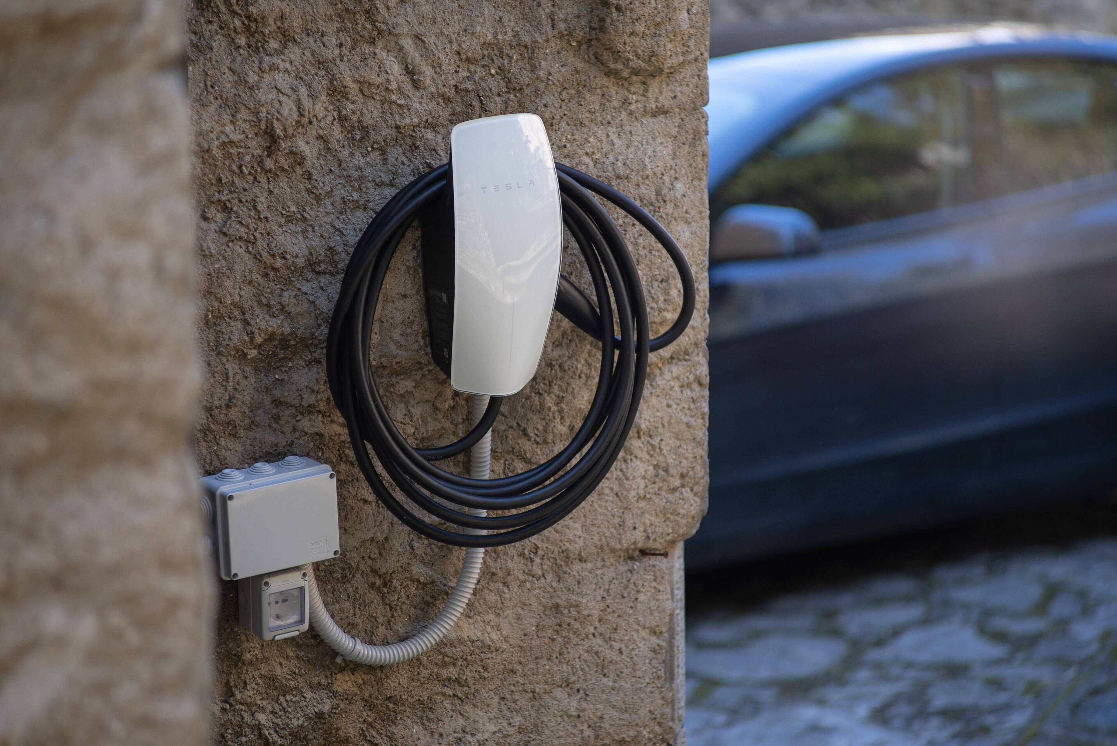 A Tesla charging station mounted on a stone wall with a car in the background.