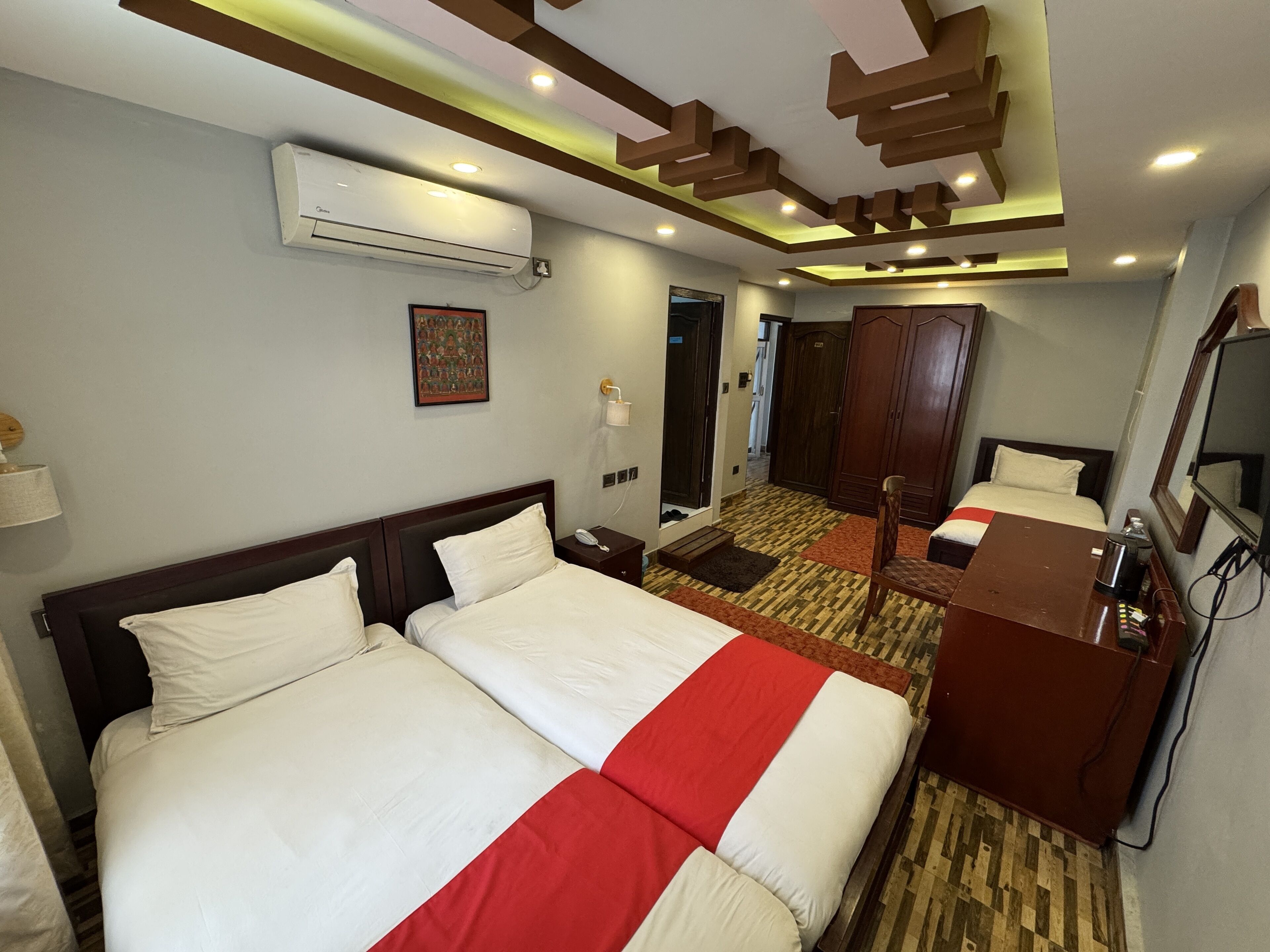 Une chambre d’hôtel avec deux lits, un bureau, un miroir, un appareil de climatisation et une porte donnant sur une autre chambre.
