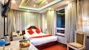 A hotel room with a bed, a desk with a basket of fruit, a chair, a window with curtains, and a TV mounted on the wall. - Hotel BnB Mhepi (Kathmandu)