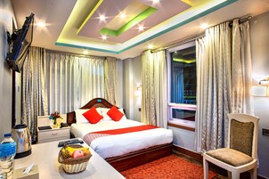 Quarto de hotel com uma cama, uma escrivaninha com uma cesta de frutas, uma cadeira, uma janela com cortinas e uma TV fixada na parede.