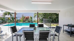 An outdoor patio with a glass table and chairs, a pool, and a view of a garden and building in the background. - Beach & Boats Agnes Water (AGNES WATER)