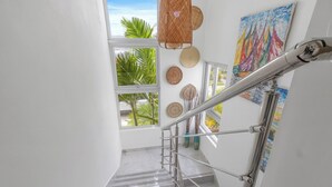 Un escalier avec une rampe en métal, des murs blancs et une vue sur des palmiers à travers de grandes fenêtres.