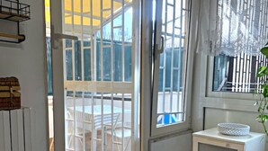 Una habitación con puerta de vidrio corredera que da a un espacio exterior con una mesa y sillas. Hay un armario blanco con cajones y un cortinilla de encaje en la ventana.