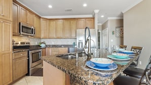 A kitchen with wooden cabinets, stainless steel appliances, granite countertops, and a dining area with chairs and a table. - Serenity Sands-Gulf Front 4 Bed Home Sleeps 10. Pup Friendly! (Navarre)