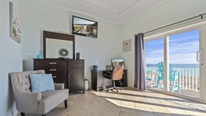 A spacious room with a large window overlooking a beach, a comfortable chair, a desk, and a television mounted on the wall. - Serenity Sands-Gulf Front 4 Bed Home Sleeps 10. Pup Friendly! (Navarre)