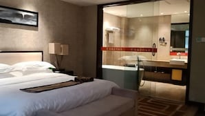 Habitación de hotel con cama, mesita de noche, lámpara y puerta de vidrio que da a un baño con ducha y espejo.