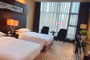 Quarto de hotel com duas camas, uma escrivaninha, uma cadeira, um vaso de planta e uma janela grande com cortinas.
