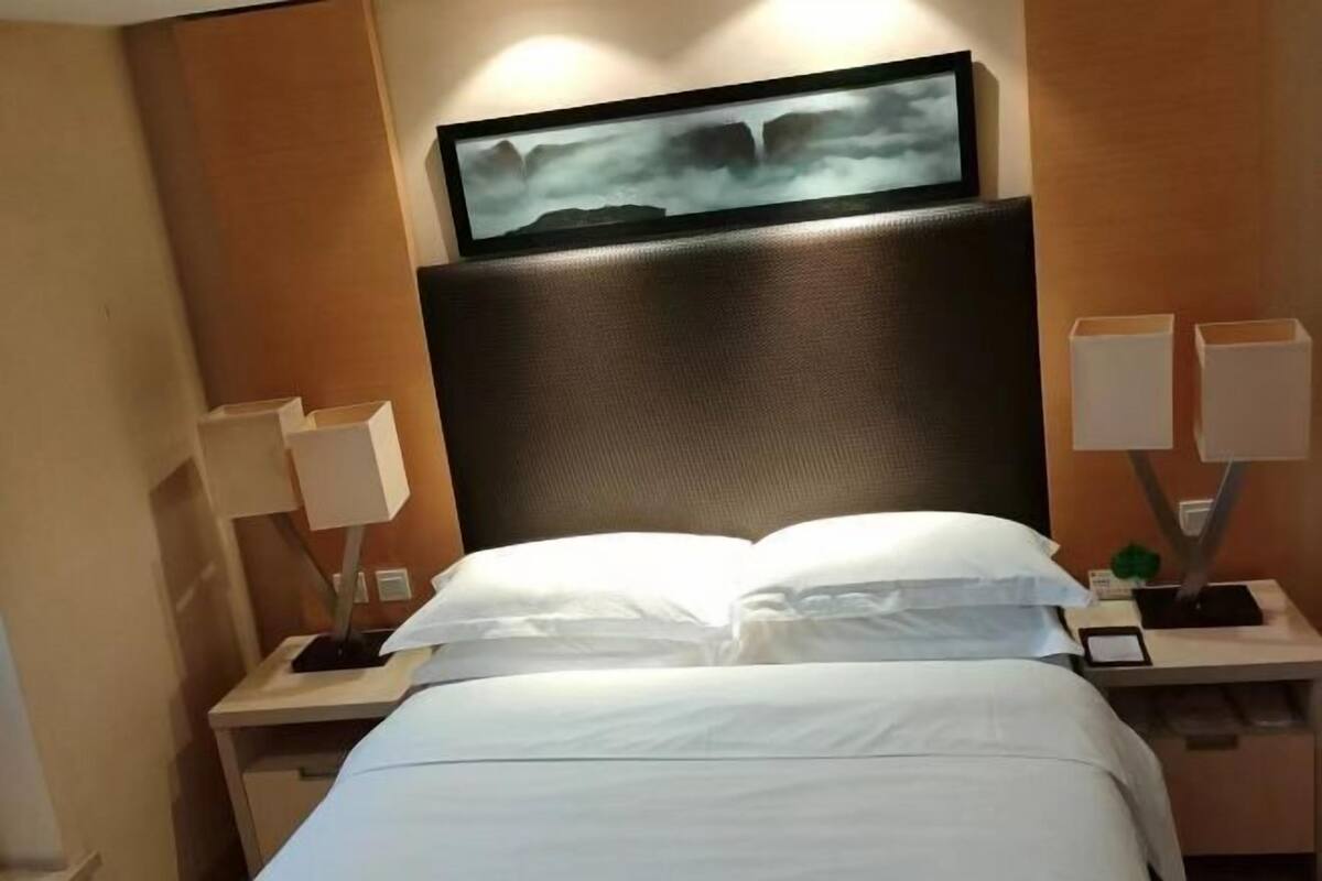 A hotel room with a bed, two bedside lamps, a framed picture above the bed, and a nightstand with a small plant.