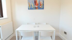 Une table à manger blanche avec quatre chaises, un tableau représentant des voiliers colorés accroché au mur et une fenêtre sur la gauche.