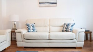 Um sofá de couro branco com duas almofadas estampadas, uma luminária e um quadro na parede.