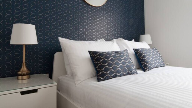 Cama bem arrumada com lençóis brancos e duas almofadas estampadas, mesa de cabeceira com abajur e parede com padrão geométrico.