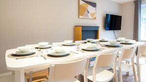 Un coin repas avec une longue table dressée pour six personnes, des chaises blanches et un téléviseur à écran plat fixé au mur.