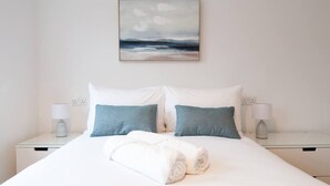 Un lit bien fait, avec des oreillers blancs et bleus, une serviette blanche roulée et, au mur, un tableau représentant un ciel nuageux au-dessus de l’eau.