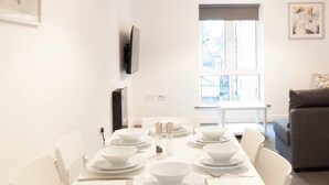 A dining area with a white table set for four, white chairs, a window with a view, and a gray sofa in the background.