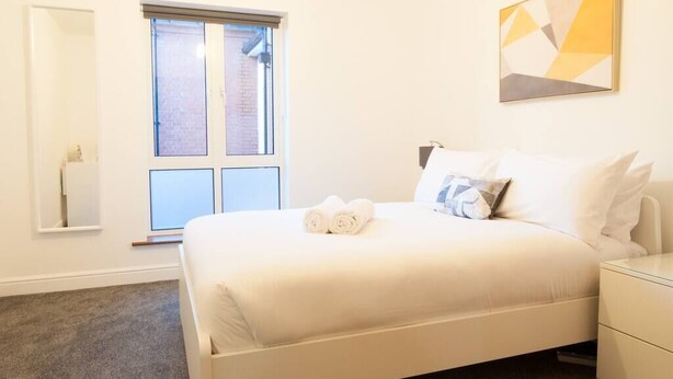 A neatly arranged bedroom with a bed, a bedside table, a window with blinds, and a wall-mounted abstract painting.