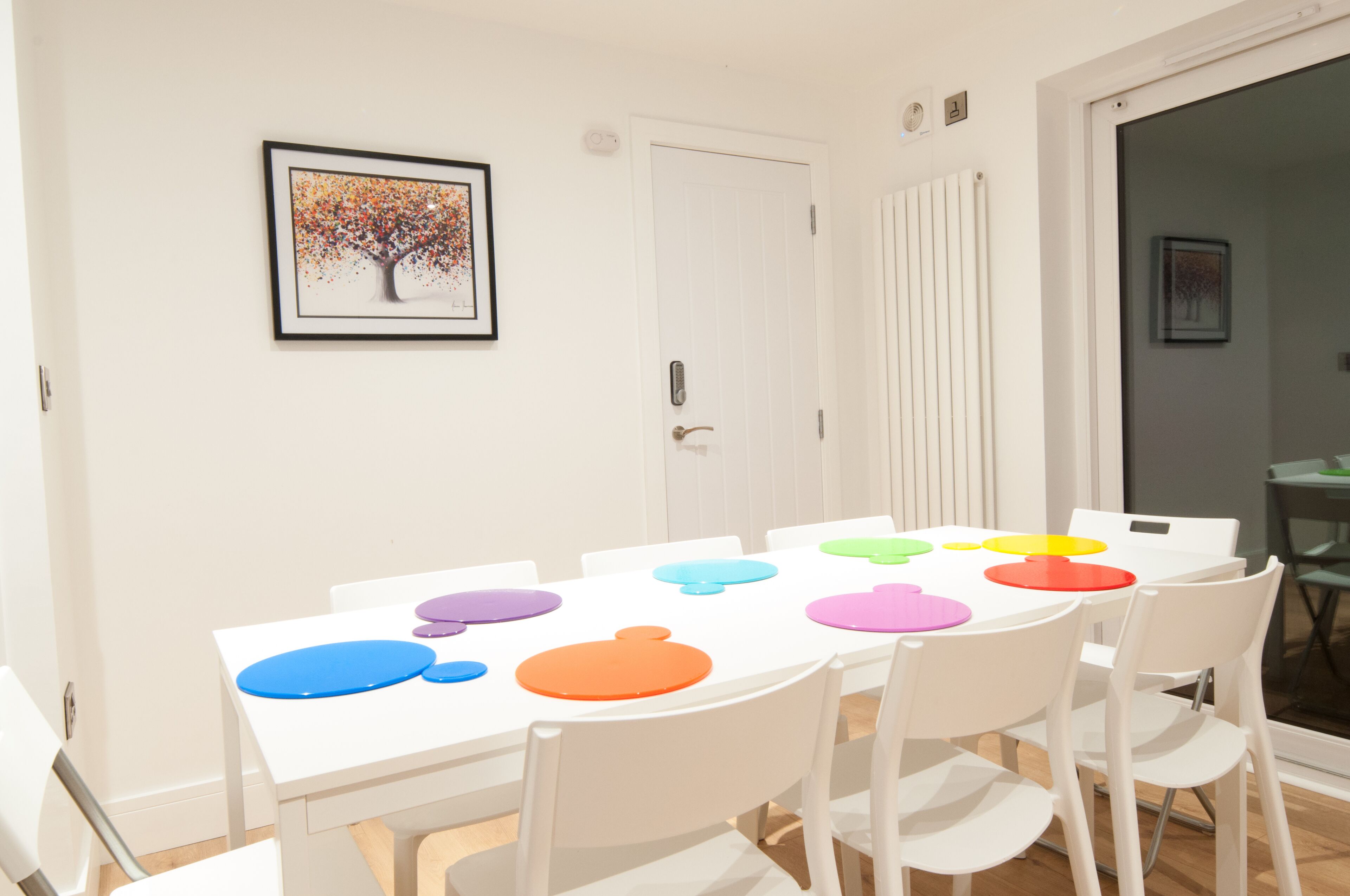 A dining area with a white table and chairs, a framed picture on the wall, and a door with a handle.