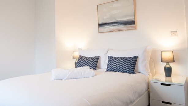 A neatly made bed with white bedding and two patterned pillows, a bedside table with a lamp, and a wall-mounted painting above the bed. - The Spitfire - Newly renovated 3-bed house in Cheltenham (Cheltenham)