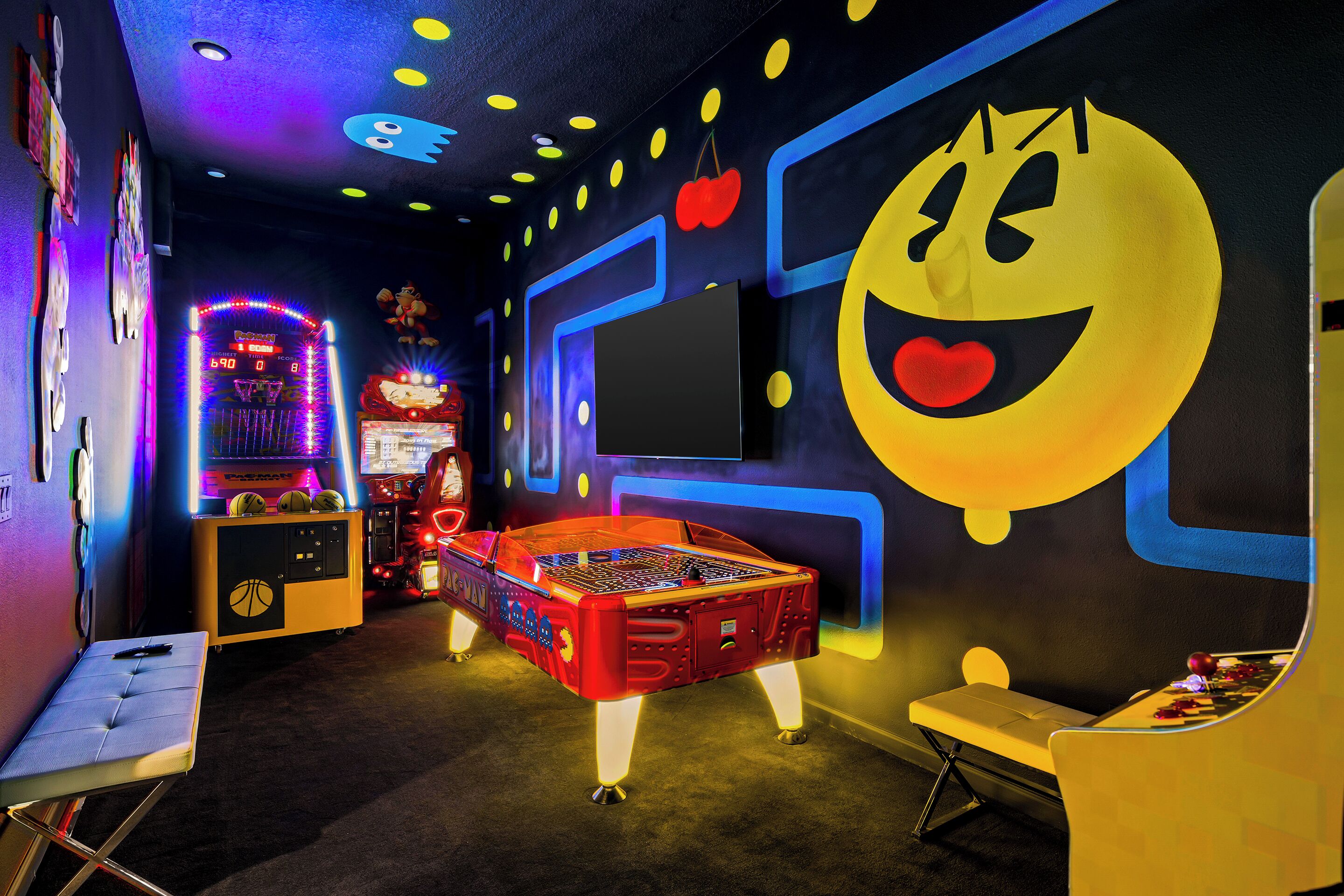 A brightly lit arcade room with neon lights, a foosball table, and a large smiley face mural on the wall.
