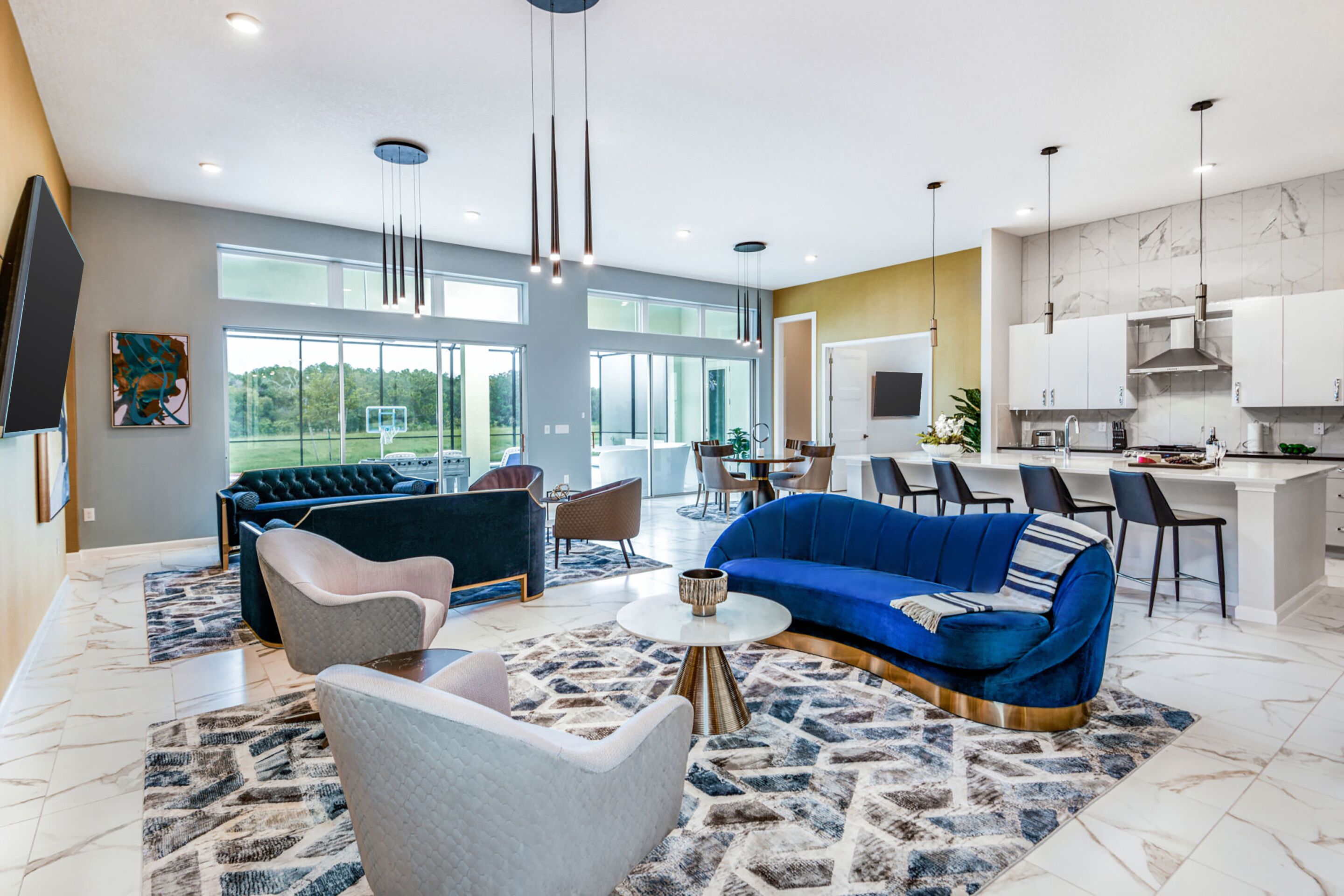 A spacious living room with a blue sofa, a grey armchair, and a coffee table. There is a dining area with a white island and chairs. The kitchen has white cabinets and a marble backsplash.