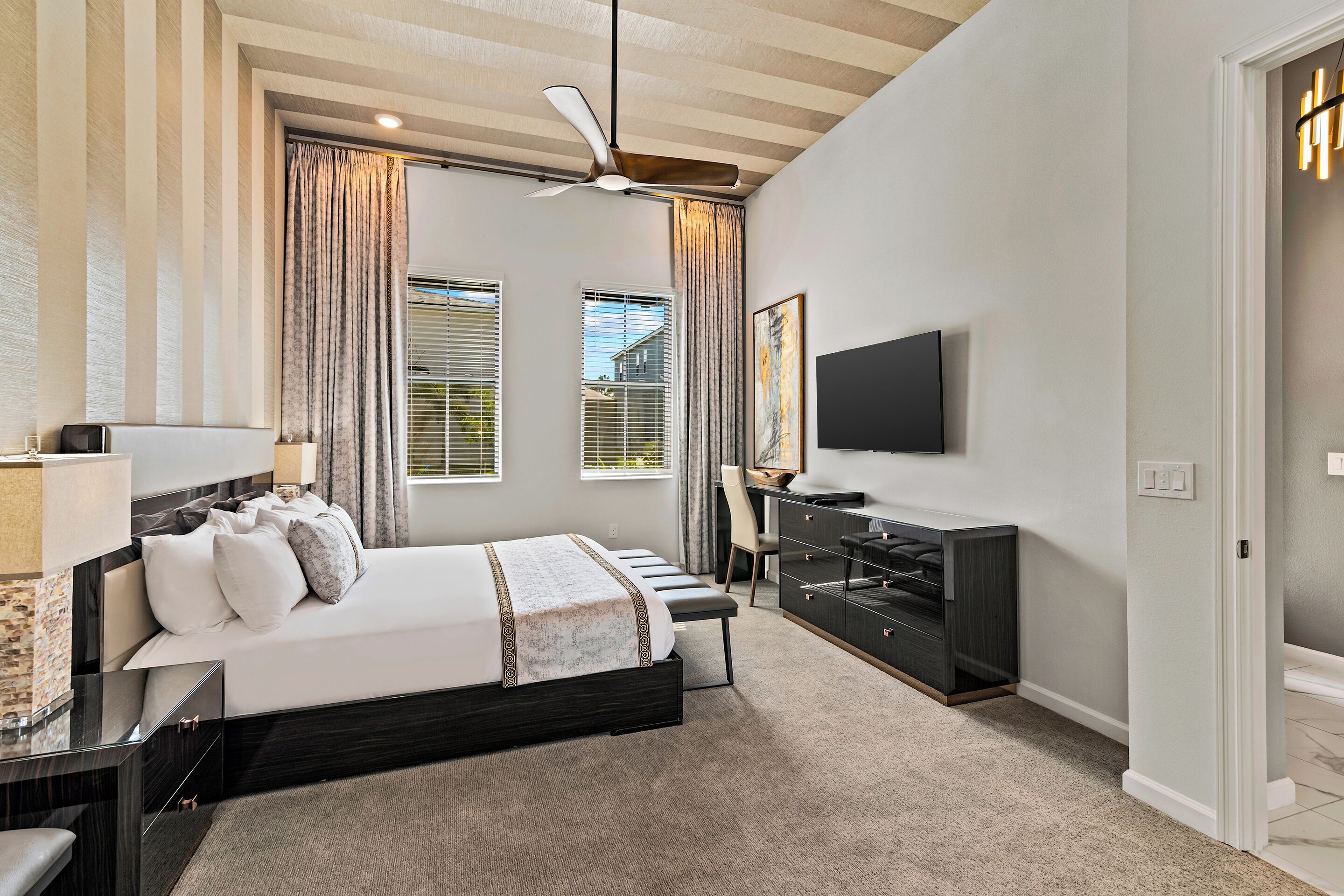 A modern bedroom with a large bed, a desk, a chair, a TV, and a ceiling fan.