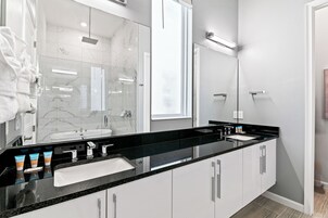 Un baño moderno con encimera negra, armarios blancos, un espejo grande y una mampara de cristal.