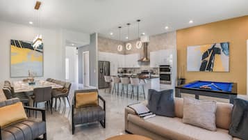 A modern living room with a pool table, a dining area, and a kitchen in the background.
