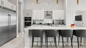 A modern kitchen with a large island, stainless steel appliances, and black bar stools. - The Village, Homes by Villatel (Davenport)
