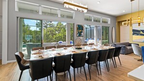 A modern dining area with a long table, black chairs, and large windows offering a view of the outdoors. - The Village, Homes by Villatel (Davenport)