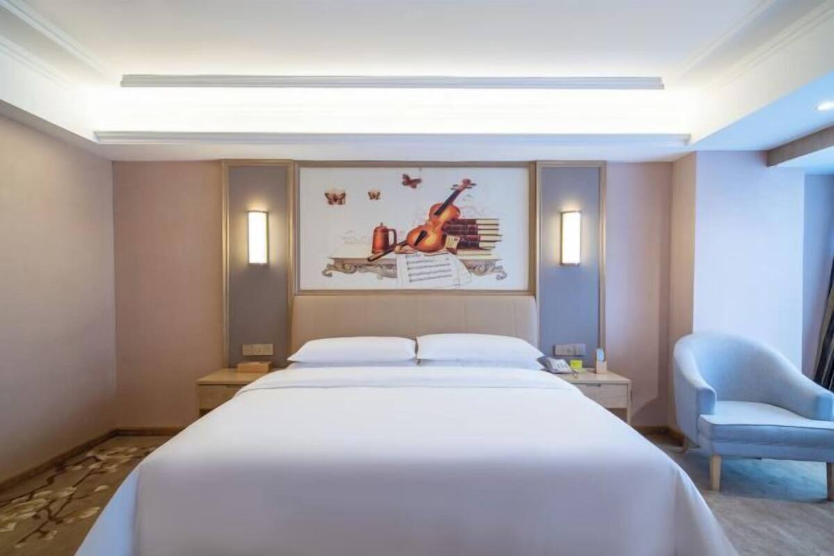 A hotel room with a large bed, a desk with a chair, and a wall-mounted artwork depicting a guitar and books.
