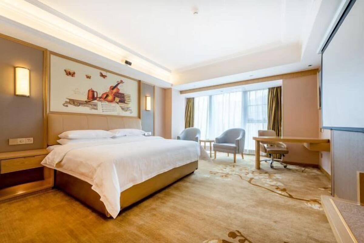 A hotel room with a large bed, a desk, two chairs, a window with curtains, and a wall-mounted artwork.