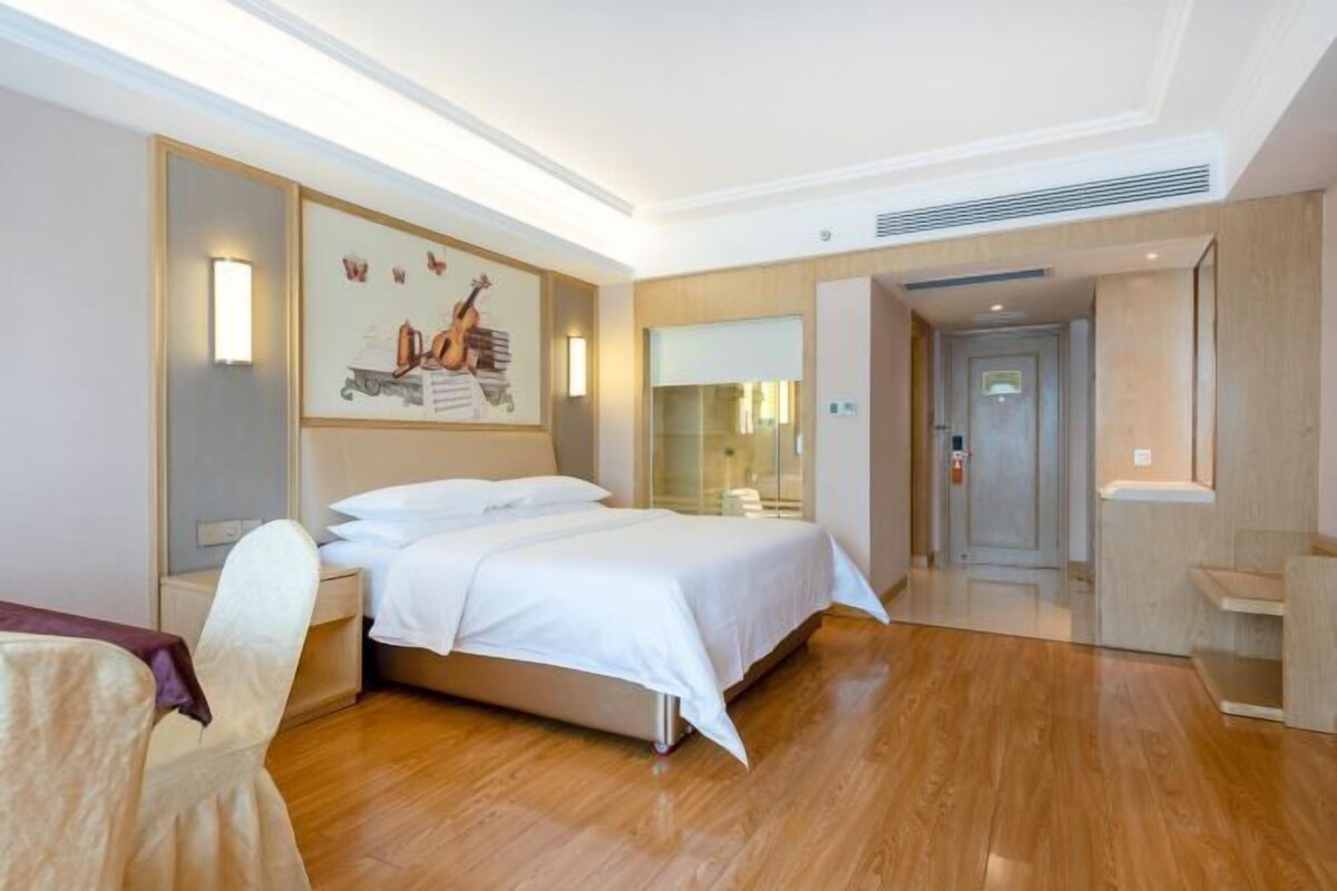 A hotel room with a bed, a desk, a chair, a painting on the wall, and a door leading to another room.