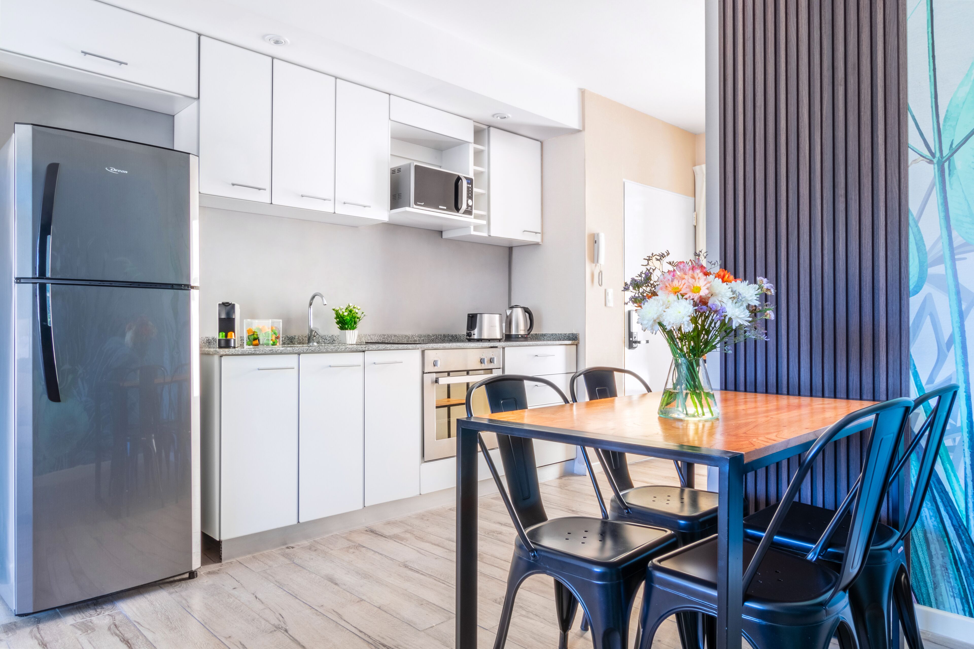 Una cocina moderna con armarios blancos, un refrigerador de acero inoxidable, un microondas y una cafetera. Hay una mesa de comedor con sillas y un florero con flores.