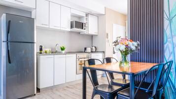 Una cocina moderna con armarios blancos, un refrigerador de acero inoxidable, un microondas y una cafetera. Hay una mesa de comedor con sillas y un florero con flores.