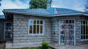 A stone house with a tiled roof, two windows with metal bars, and a small bush in the front yard.