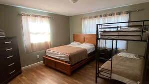 Een kamer met een houten vloer, een enkel bed met een houten hoofdbord, een stapelbed, een dressoir en een raam met gordijnen.