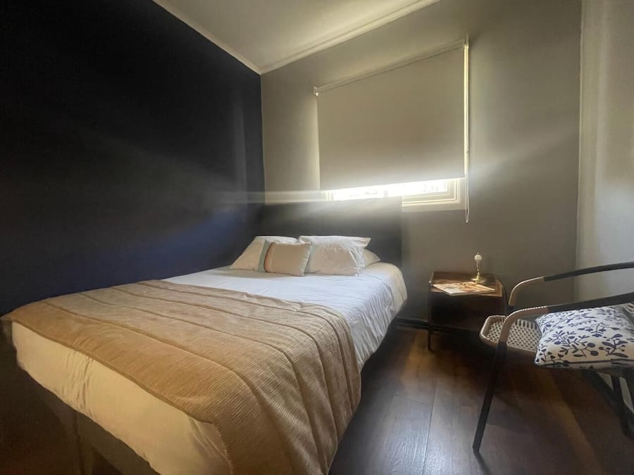 Quarto com cama, mesa de cabeceira, cadeira e janela com persianas.