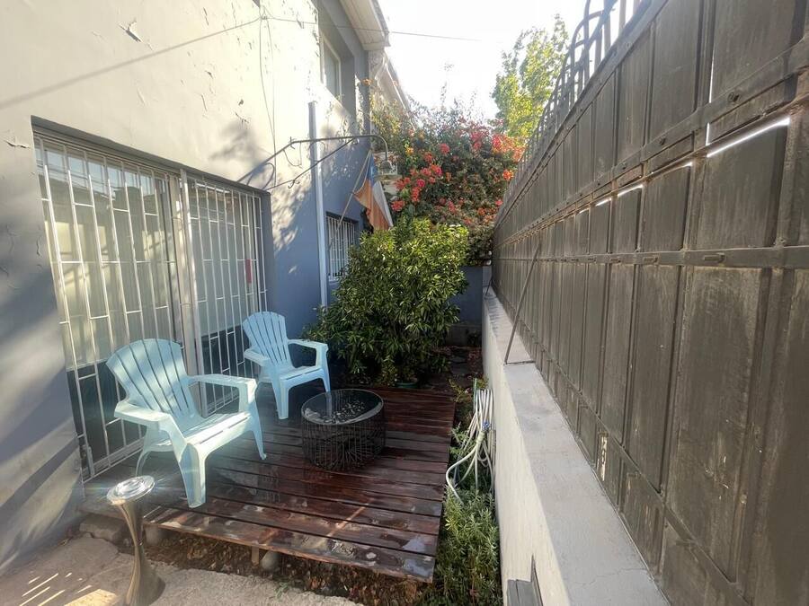 Un pequeño patio con dos sillas blancas y una mesa pequeña, rodeado por una terraza de madera y una cerca de metal.